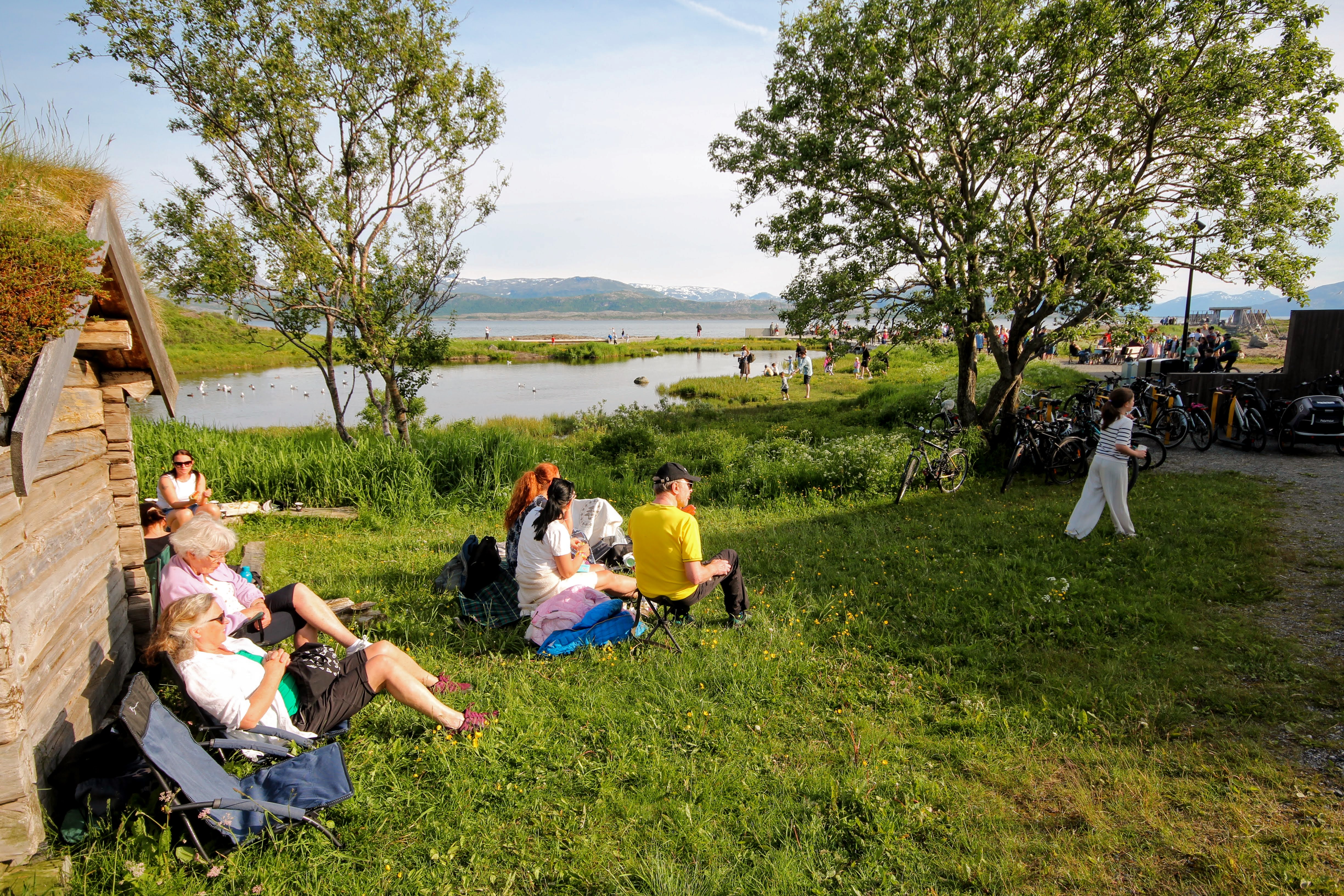 Noen mennesker sitter i solveggen ved et laftet hus med torvtak. Ei jente går på gresset. Det er vann og fjell i bakgrunnen. Noen bjørketrær på begge sider.